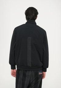 Crna jakna s visokim ovratnikom, teksturirana tkanina i tri vertikalne pruge na leđima. Manžetne i rub su elastični za udoban fit.