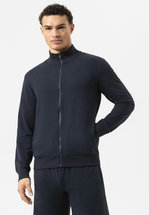 Man draagt een marineblauw jack met rits en hoge kraag en bijpassende broek, staat met één hand in de zak tegen een effen achtergrond.