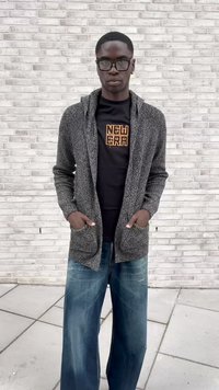 Ung mand iført sorte briller, grå cardigan, sort "New Era" t-shirt og brede blå jeans, der står op ad en hvid murstenvæg.