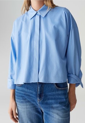 Femme portant une chemise ample bleu clair à boutons avec des manches longues associée à un jean en denim bleu sur un fond neutre.