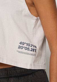 Jasnoszary bawełniany top na ramiączkach o kroju cropped. Zawiera czarny tekst z współrzędnymi oraz frazą po lewej stronie. Miękka tekstura.