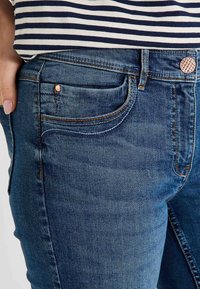Jeans en denim bleu foncé avec une texture lisse, présentant un design classique à 5 poches et un bouton métallique décoratif. Légère décoloration sur les cuisses.