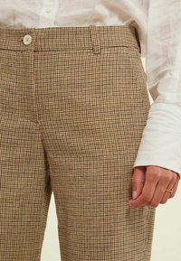 Pantalons à carreaux marron en tissu texturé, avec une fermeture éclair et un bouton. Les poches latérales sont visibles, avec une coupe ajustée.