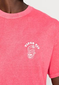 Mörkhyad person som bär en rosa T-shirt med en vit "VIPER PIT" logotyp som visar en sammanflätad orm och ett skalle på bröstet.