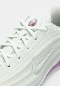 Λευκό αθλητικό παπούτσι με υφασμάτινο επάνω μέρος με υφή, γκρι λεπτομέρειες, μώβ λεπτομέρεια στη σόλα και λευκό λογότυπο Nike στο πλάι.