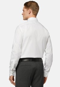 Camicia bianca a maniche lunghe con colletto classico, tessuto liscio e polsini con bottoni, abbinata a pantaloni grigi scuri e una cintura nera.