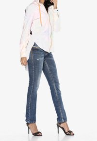 Chaqueta blanca iridiscente con cremallera y cordón naranja, combinada con jeans de mezclilla azules que presentan acentos desgastados y costura lateral en contraste.