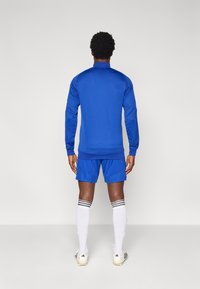 Μπλε αθλητική φόρμα με μακρυμάνικο τοπ και σορτς. Διαθέτει ριμπ μανσέτες και τελείωμα, συνδυασμένη με λευκές κάλτσες και ποδοσφαιρικά παπούτσια.