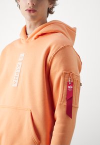 Sudadera naranja con un bolsillo delantero, que presenta un logotipo blanco "ALPHA" y un bolsillo lateral con una cremallera roja y detalles de etiqueta. Textura de tela suave.