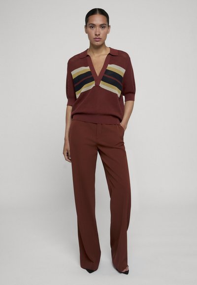 Femme debout portant un pantalon couleur rouille et un haut en maille à manches courtes couleur rouille avec des rayures noires, jaunes et beiges traversant la poitrine.