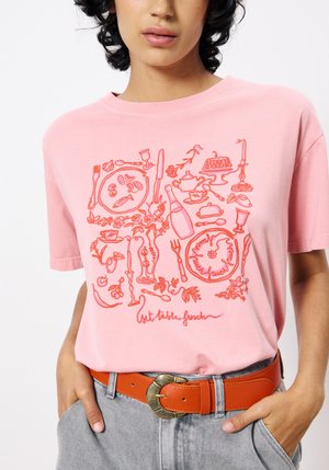 Rosa Baumwoll-T-Shirt mit einem roten, lebensmittelthemen Grafikdesign. Kurze Ärmel, runder Ausschnitt, kombiniert mit einem orangefarbenen Gürtel und grauen Hosen.