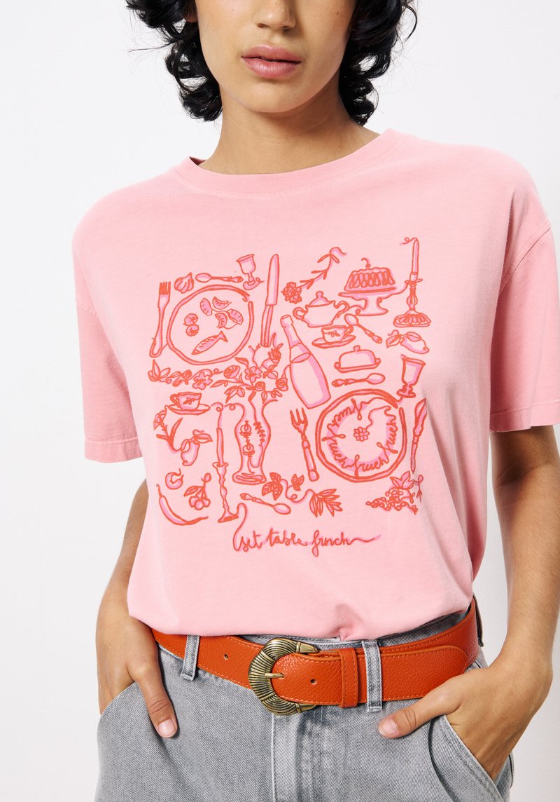 Roze katoenen t-shirt met een rood voedselthema grafisch ontwerp. Korte mouwen, ronde halslijn, gecombineerd met een oranje riem en grijze broek.