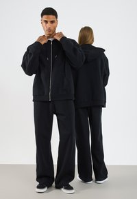 Svart zip-up hettegenser med snor og ribbestrikkede mansjetter, kombinert med matchende løse bukser. Begge har en myk tekstur og minimalistisk design.