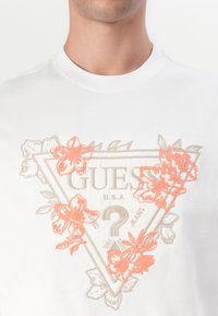 Bílý bavlněný tričko s trojúhelníkovým designem a béžovou a korálovou květinovou výšivkou, uprostřed s logem "GUESS U.S.A."