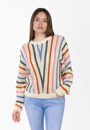 Femme portant un pull en maille crème à rayures verticales orange, vert, rose et bleu, associé à un jean bleu clair, debout sur un fond blanc.