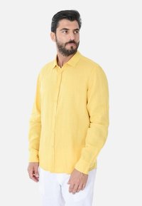 Camicia gialla di lino a maniche lunghe con abbottonatura frontale, colletto standard e maniche arrotolate. Indossata con pantaloni bianchi.