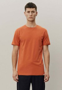 Kortermet oransje T-skjorte laget av bomull med rund hals og en liten brodert detalj på brystet, med en glatt tekstur.