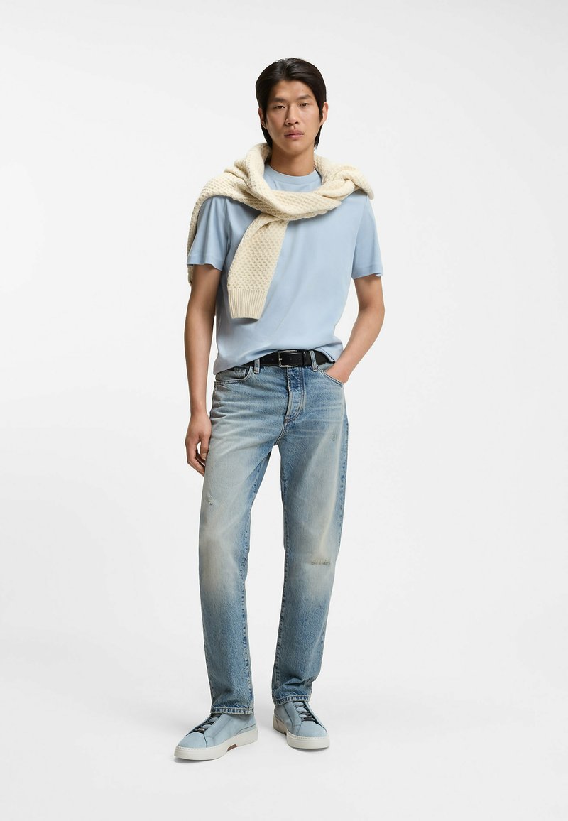 Giovane uomo in piedi con una mano in tasca, indossa una maglietta azzurro chiaro, jeans sbiaditi, sneakers blu e un maglione crema di maglia appoggiato sulle spalle.