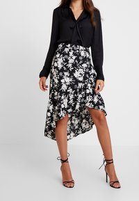 Mustat puserot, jossa on nauha kaulassa, yhdistettynä musta-valkoiseen kukkakuosisena epäsymmetriseen hameeseen, jossa on rypytyksiä. Mustat nauhalla varustetut korkokengät.