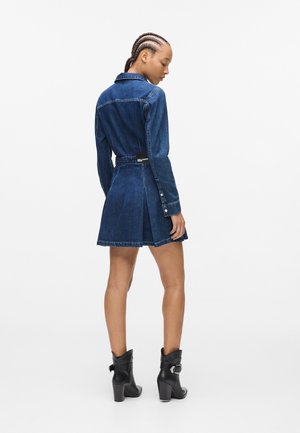 Denim-Hemdkleid mit langen Ärmeln, dunkler Waschung, tailliertem Schnitt, ausgestelltem Rock, Knopfverschluss und einem Schlitz auf der Rückseite. Schwarze Knöchelstiefel runden den Look ab.