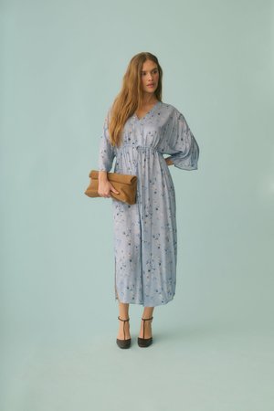 Kobieta w niebieskiej sukience midi w kwiaty z bufiastymi rękawami trzyma brązową kopertówkę, nosi czarne czółenka na pasku w kształcie litery T, stoi na jednolitym jasnoniebieskim tle.