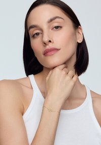 Złota bransoletka z okrągłym zawieszeniem, noszona na ręce spoczywającej na podbródku. Modelka ma na sobie prosty biały top na ramiączkach, a tło jest neutralne.