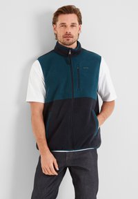 Man standing against a wall wearing a two-tone blue fleece vest over a white short-sleeve shirt and dark jeans, hands in pockets.