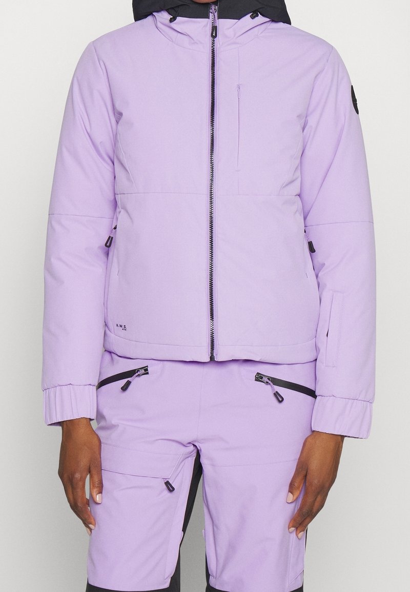 Personne portant une veste violet clair avec une fermeture éclair noire et un pantalon assorti avec plusieurs poches zippées, debout devant un fond uni.