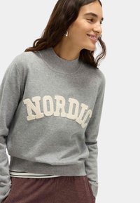 Grijze sweatshirt met hoge hals, met "NORDIC" in crèmewit, textuurstof, ribgebreide manchetten en tailleband, gecombineerd met donkere broeken.