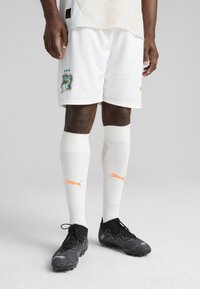 Shorts de foot blancs avec un blason ivoirien vert et orange, associés à des chaussettes blanches avec un logo Puma orange et des crampons de foot noirs.
