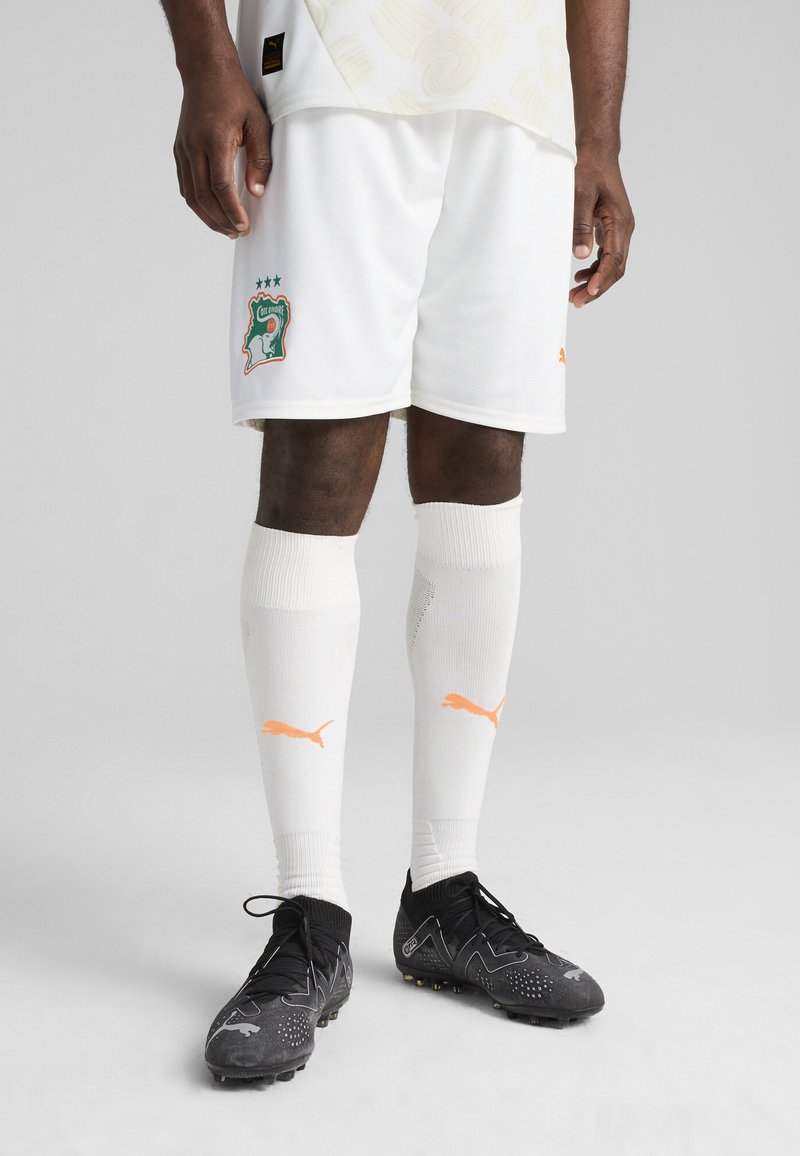 Hvide fodboldshorts med grøn og orange ivoriansk crest, parret med hvide sokker med orange Puma-logo og sorte fodboldstøvler.