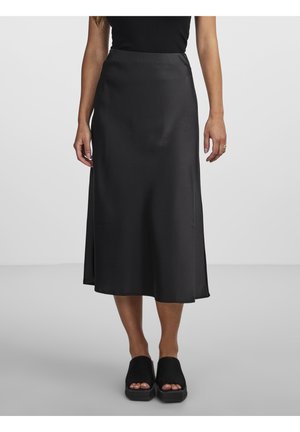Femme portant un haut noir sans manches, une jupe midi en satin noire et des sandales à plateforme ouvertes aux orteils noires, debout sur un sol gris uni.