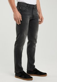 Férfi elhalványult fekete slim-fit farmerben és fekete sportcipőben barna talppal, semleges háttér előtt állva.