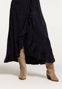Langes, schwarzes Kleid mit einem Rüschensaum und subtil strukturiertem Muster, kombiniert mit sandfarbenen Wildleder-Stiefeln im Cowboy-Stil mit dekorativer Naht und Blockabsatz.