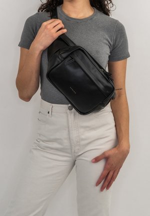 Persona che indossa una maglietta grigia e pantaloni bianchi, con una borsa a tracolla in pelle nera portata sul petto.