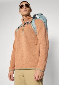 Pullover in pile arancione con collo a zip, dettagli grigi e un motivo testurizzato. Indossato con uno zaino azzurro chiaro e occhiali da sole.