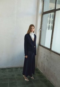 Námornícky sveter s jedným zlatým gombíkom, v kombinácii s dlhou námorníckou sukňou a bielym šatkou. Model má na sebe biele sandále na vysokom opätku.