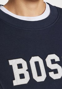 Gros plan d'une personne portant un sweat-shirt bleu marine avec des lettres blanches formant "BOSS" et un t-shirt blanc à col rond en dessous.