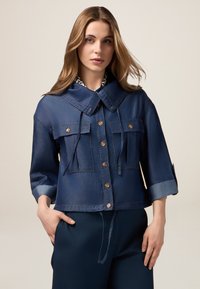 Chaqueta de mezclilla recortada en azul oscuro con gran cuello, dos bolsillos en el pecho y cierre de botón. Incluye mangas enrollables y cordón en la cintura.