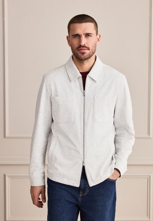 Mann mit kurzen Haaren und Bart, der eine hellgraue Reißverschlussjacke über einem dunklen Hemd trägt, steht mit einer Hand in der Tasche vor einem einfarbigen Hintergrund.