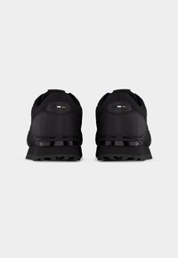 Paire de chaussures de sport noires vues de dos avec tissu texturé et petit logo rectangulaire près du talon sur un fond uni.