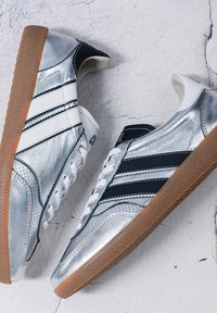Paire de chaussures de sport argentées avec des rayures noires et blanches et des semelles en caoutchouc sur une surface blanche fissurée.
