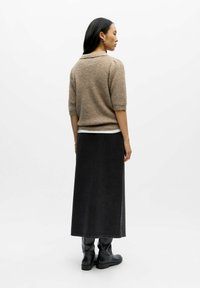 Donna con capelli lunghi e scuri che sta in posizione laterale, indossando un maglione corto marrone a maniche corte, una gonna midi nera e stivali neri su sfondo bianco.