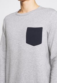 Camisa de manga larga gris de algodón, con un bolsillo azul marino en el lado izquierdo del pecho, de cuello redondo y con una textura sutil.