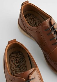 Chaussures en cuir marron avec détails en brogue, bout rond et design à lacets. L'intérieur présente une semelle en texturé avec un motif en vagues.