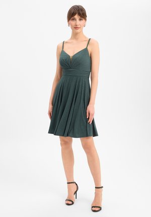 Frau steht und trägt ein ärmelloses, dunkelgrünes knielanges Kleid mit plissiertem Oberteil und schwarze, hochhackige Sandalen mit Knöchelriemen.