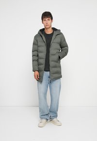 Πουφ μποστάνι μπουφάν σε σκούρο πράσινο χρώμα, με κουκούλα και κατακόρυφες ραφές. Συνδυάζεται με μαύρο πουκάμισο και ανοιχτό μπλε τζιν, παρέα με άσπρα sneakers.