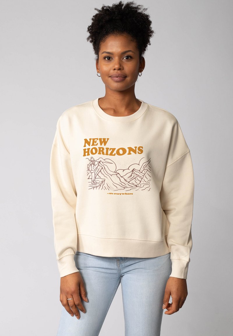 Crème kleurige oversized sweatshirt met "NEW HORIZONS" in oranje gedrukt. Heeft een bergafbeelding en een ontspannen halslijn. Zachte textuur.