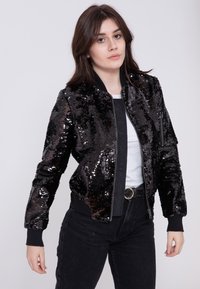 Czarna kurtka bomberka pokryta cekinami, z prążkowanym kołnierzem i mankietami, zamek błyskawiczny oraz boczne kieszenie. W zestawie z szarym t-shirtem i ciemnymi spodniami.