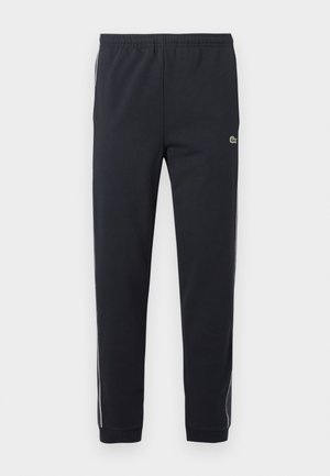 Pantalon de sport noir avec taille élastique, jambes fuselées, bandes latérales blanches et petit logo crocodile vert sur la cuisse droite.